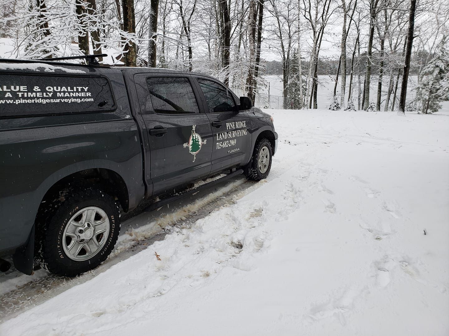 A dark gray pickup truck with business decals parked on a snow-covered road surrounded by snowy trees.
