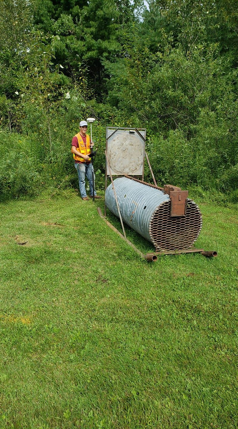 A person in a reflective vest holds a surveying pole next to a metal culvert pipe in a grassy, wooded area.