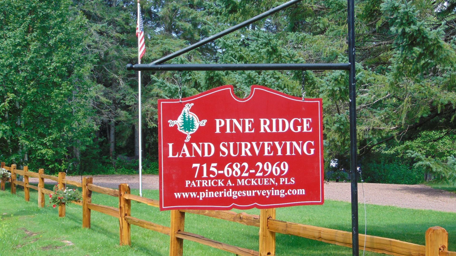 A red sign for Pine Ridge Land Surveying hangs on a black post in front of a wooden fence and trees.