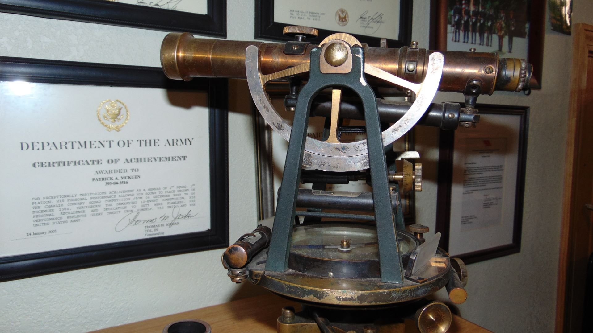 A vintage brass surveyor’s transit instrument sits on a surface, centered in front of framed documents on a wall.