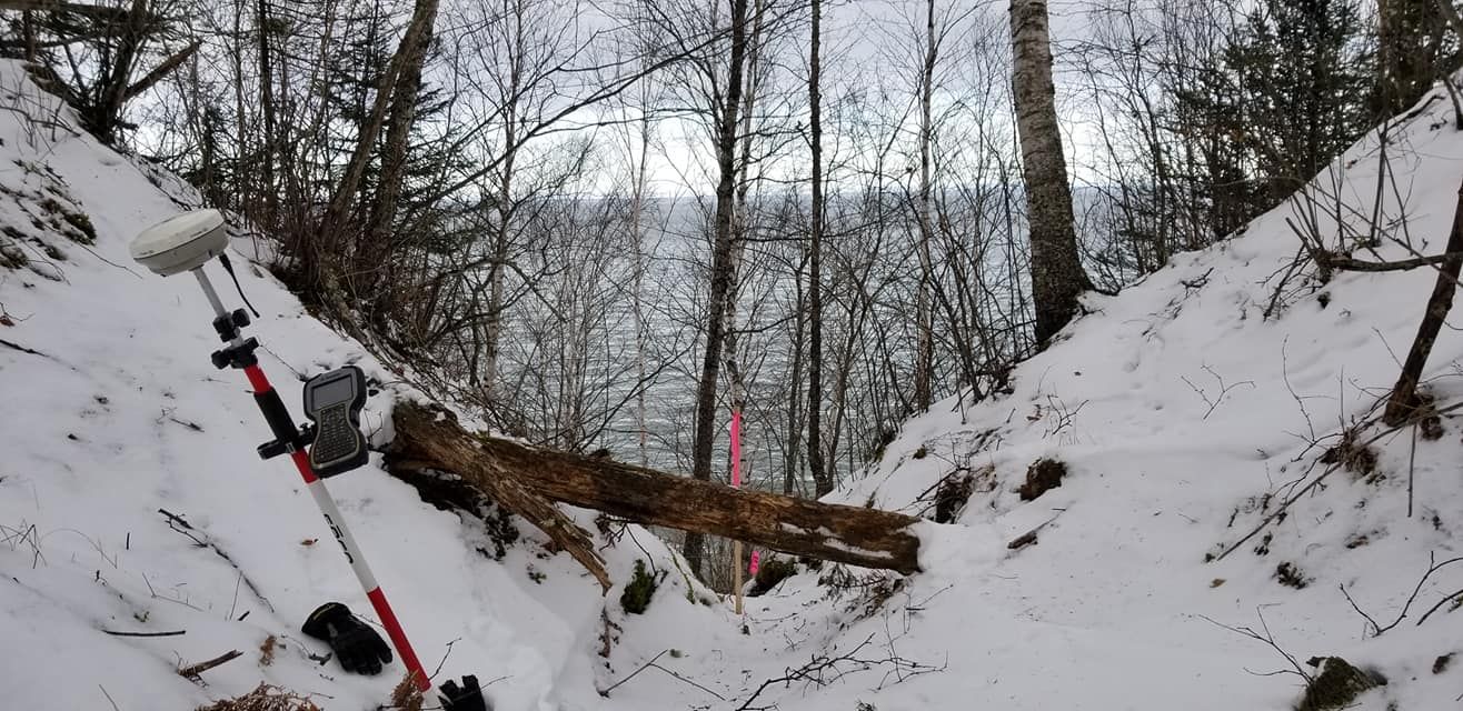 A survey pole with a GNSS receiver stands in a snowy, wooded ravine near a water body, with a fallen tree across the path.