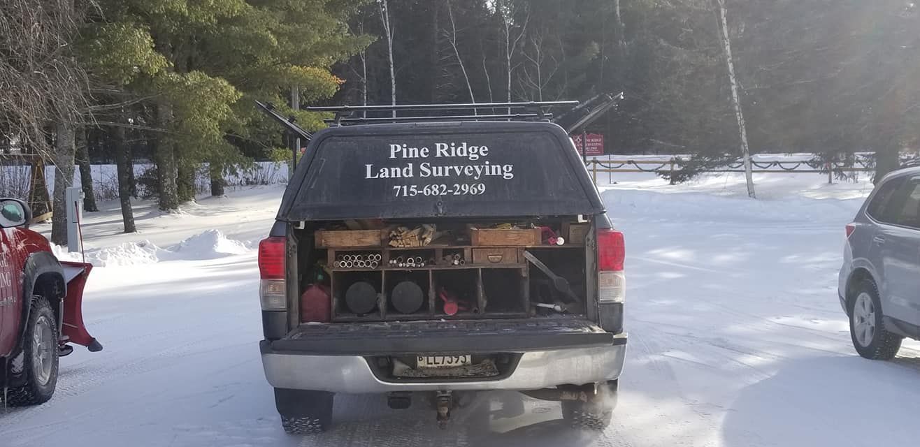 A black Pine Ridge Land Surveying truck parked in a snowy area, seen from the rear with the tailgate lowered.