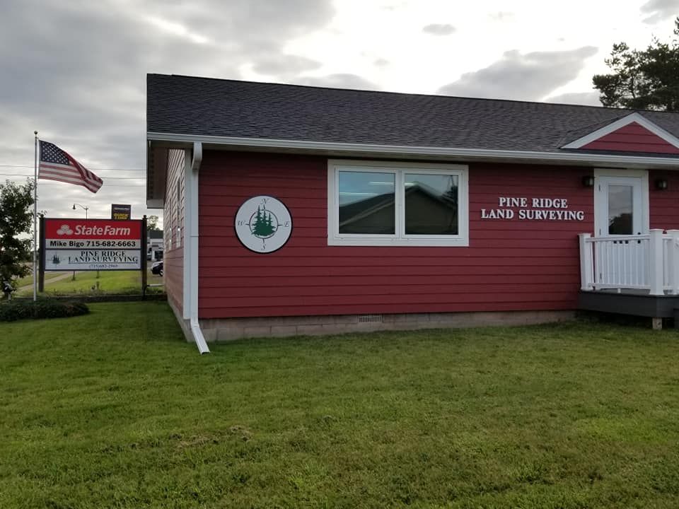 A red-sided office building for Pine Ridge Land Surveying with a lawn, a flagpole, and a State Farm sign nearby.