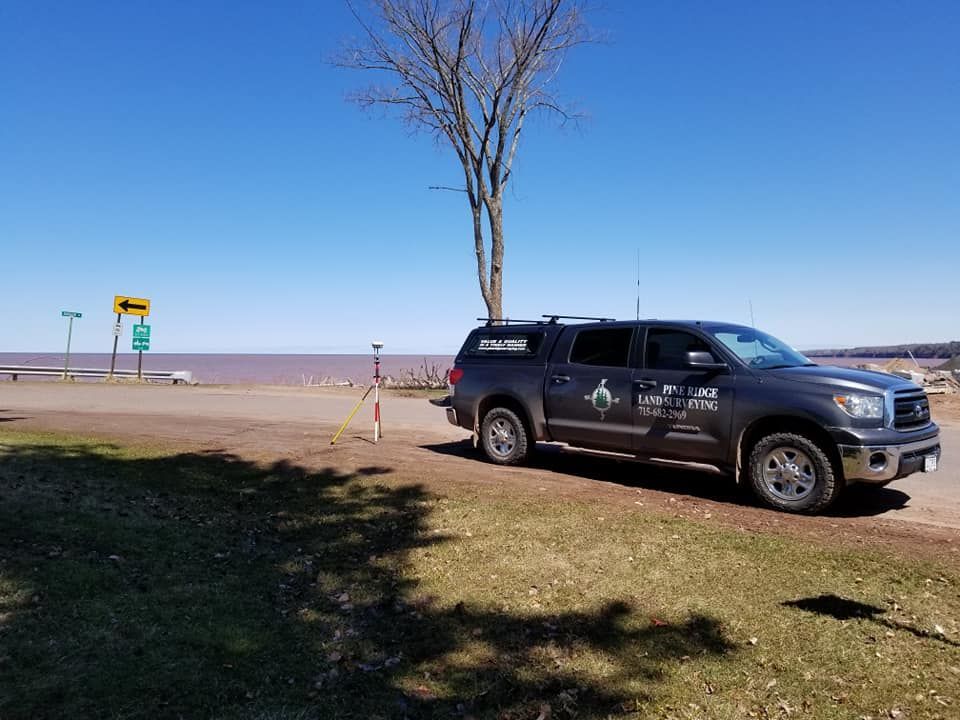 A dark gray survey truck parked on grass near a lake with a survey rod and road signs in the background.