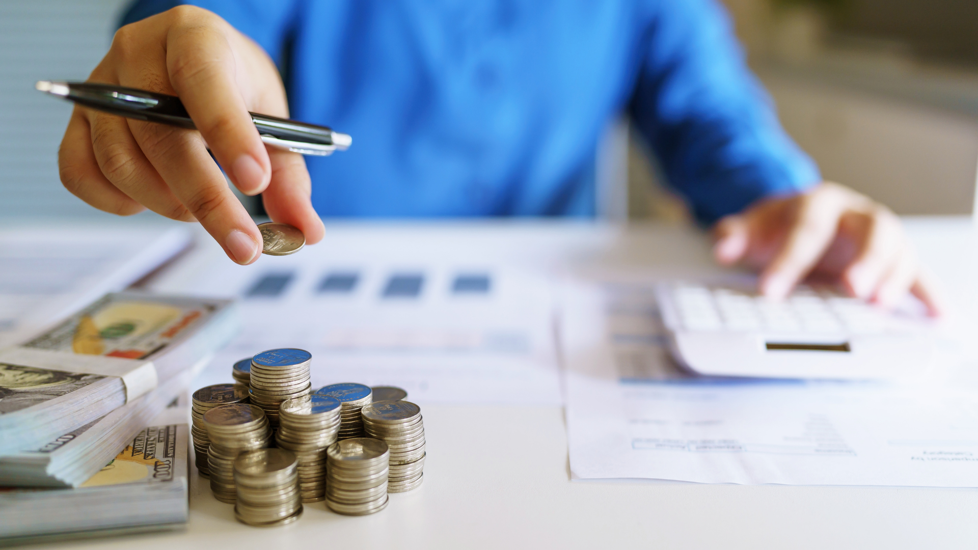 A person is holding a pen over a pile of coins while using a calculator.