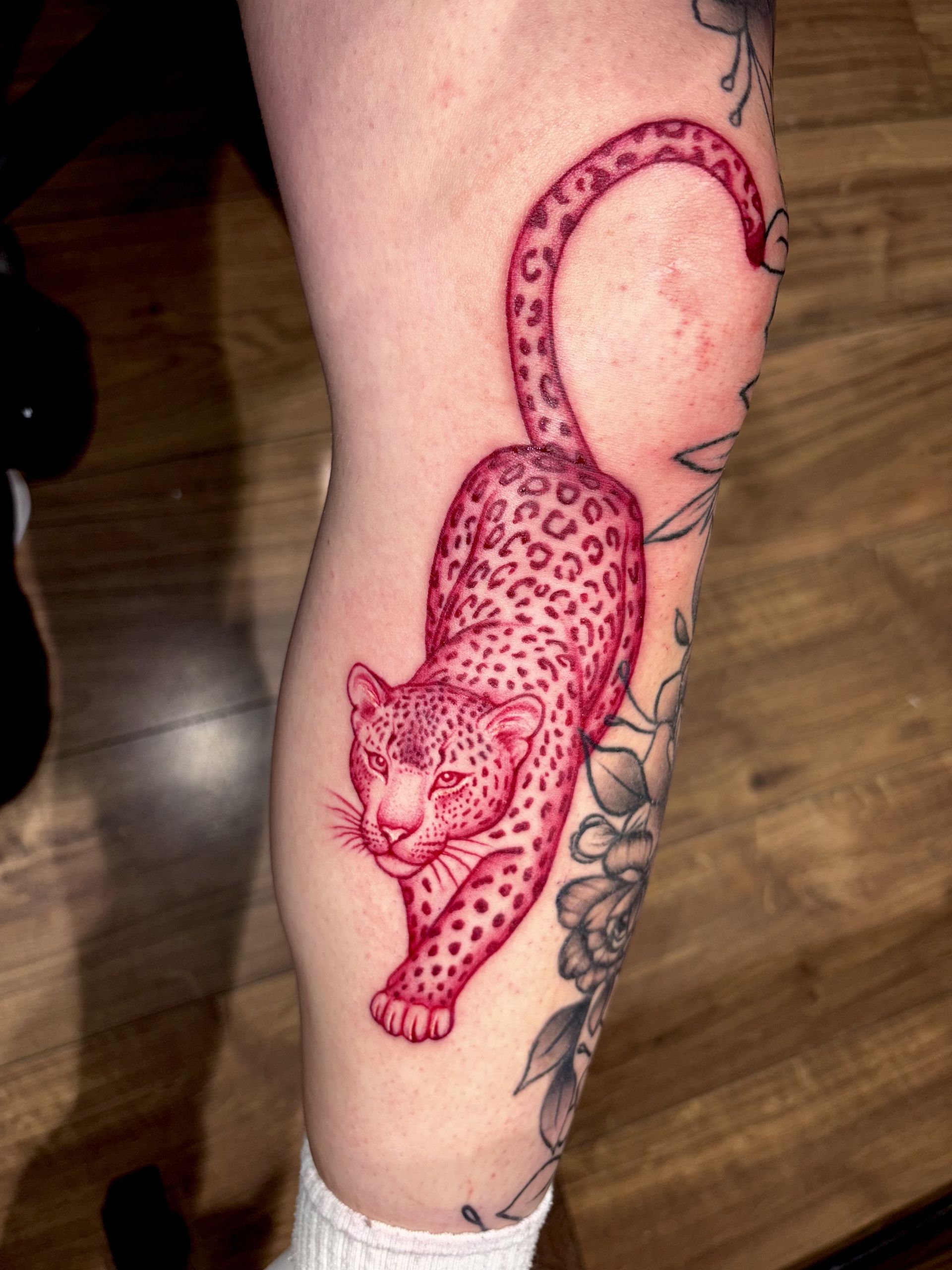 A red ink tattoo of a leopard crawling downward on a person's leg, positioned next to a black-and-grey floral tattoo.