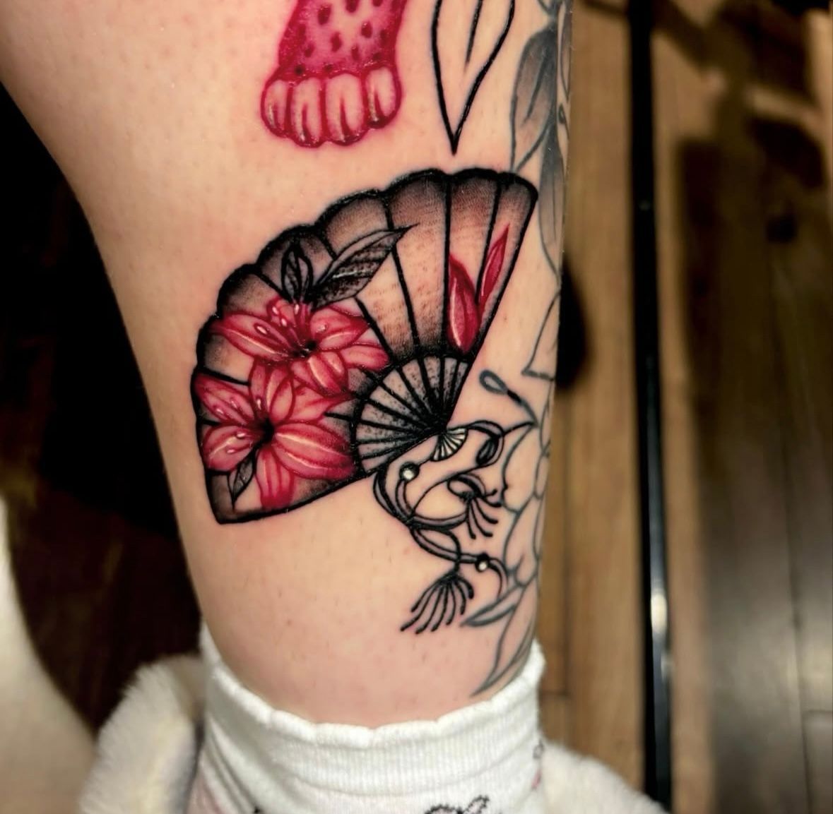 A tattoo of an open Japanese folding fan featuring red lilies and black shading on a person's leg.