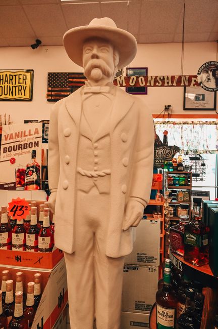 Statue of a man in a hat and suit stands inside a liquor store. Surrounded by bottles and signs.