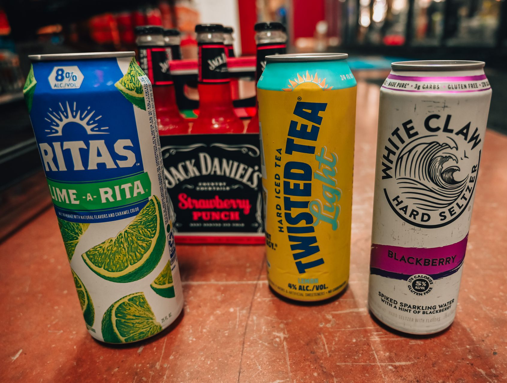 Assorted alcoholic beverages on a wooden surface: Rita's lime, Jack Daniel's Strawberry Punch, Twisted Tea Light, White Claw Blackberry.