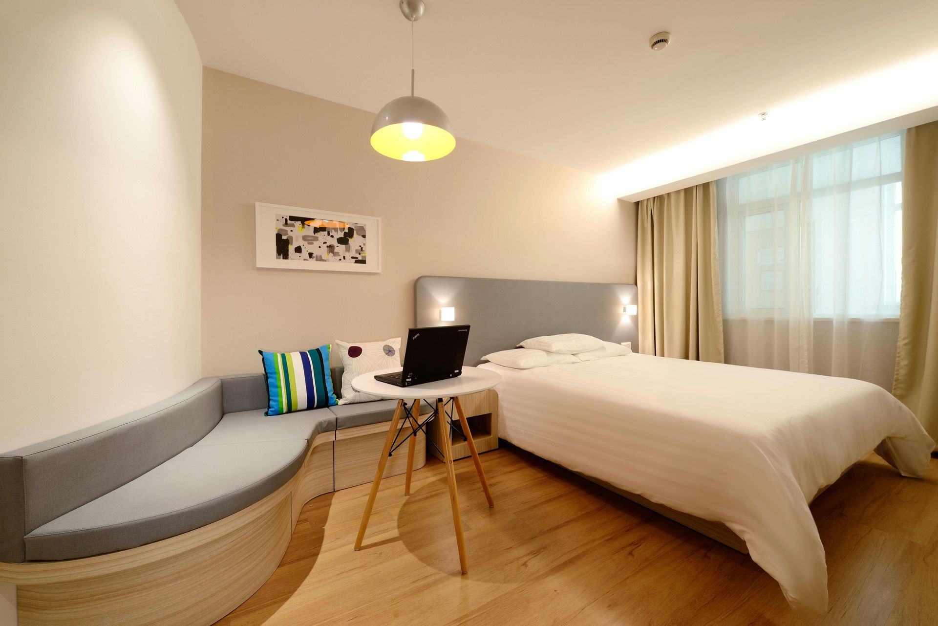 A modern hotel room with a bed, a gray curved bench, a small round table with a laptop, and beige walls.