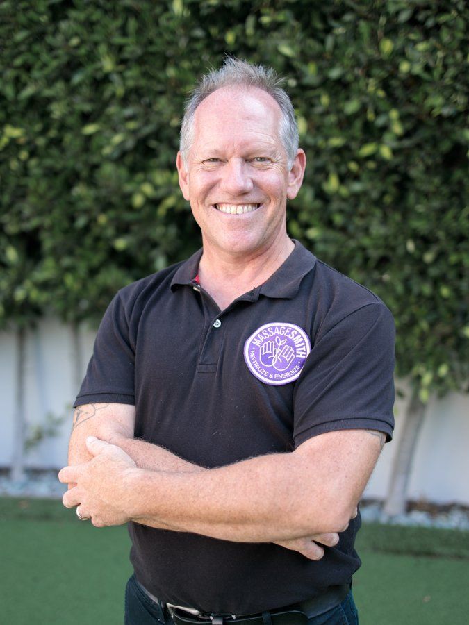 A man in a black shirt with a purple logo on it is standing with his arms crossed and smiling.