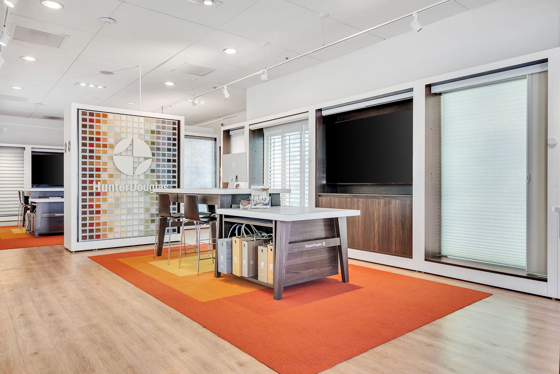 A bright, modern showroom featuring a center island, fabric swatch display, and window blind samples on a warm-toned rug.