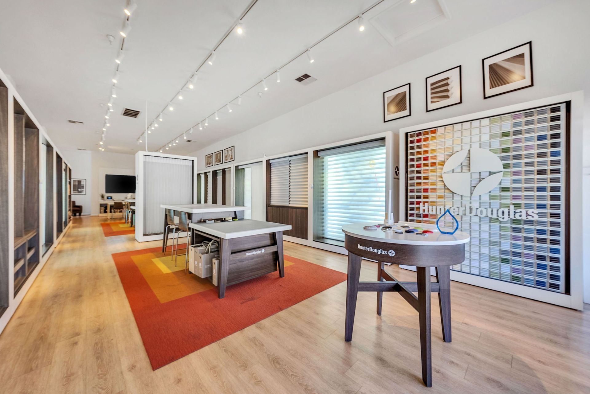 A bright, modern showroom featuring wood floors, orange area rugs, display tables, and a wall of colorful fabric swatches.