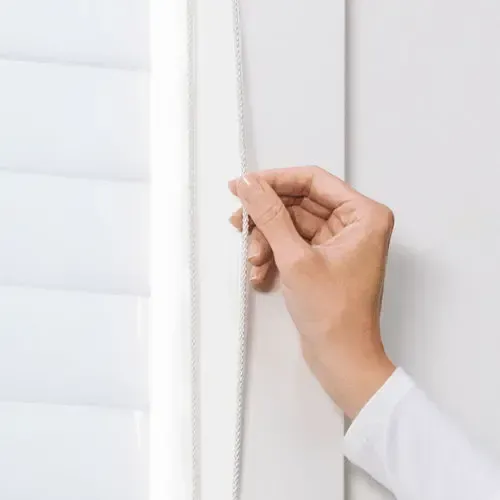 Hand pulling a cord to adjust white window shutters in a bright room.
