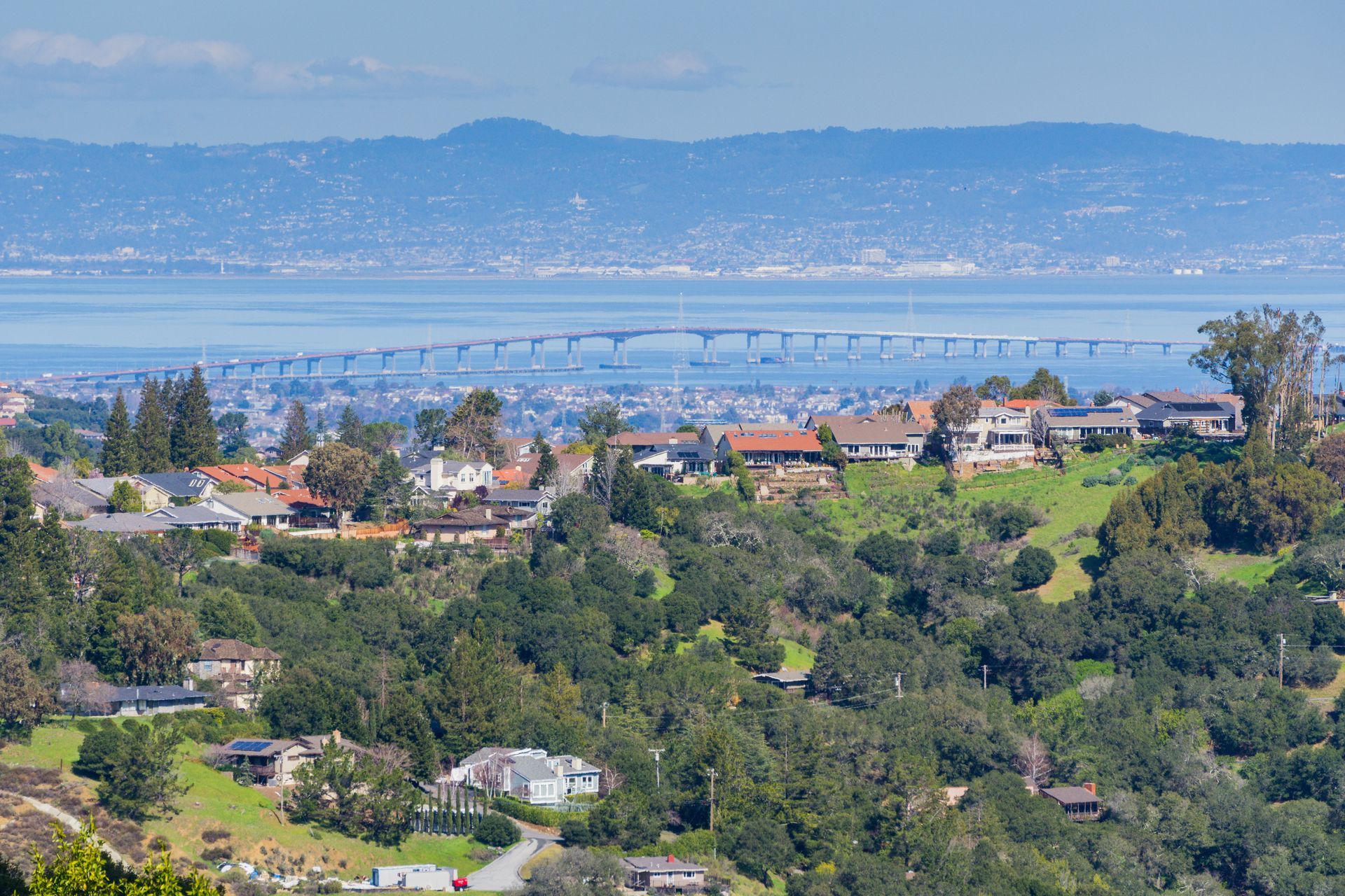 Houses on a hill overlooking a bay with a bridge and mountains in the distance on a sunny day.