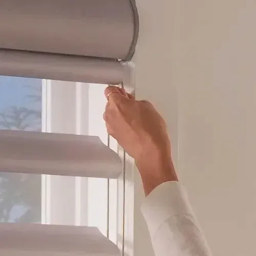 Hand adjusting light-filtering window blinds. Gray blinds with a visible control cord and white wall.