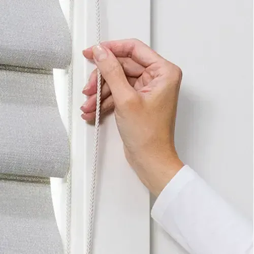 Person's hand pulling a cord to adjust a light gray Roman shade on a white window frame.