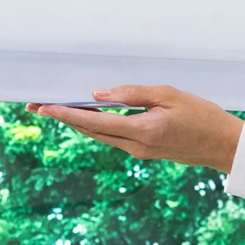 Hand adjusting a white roller shade in front of a window overlooking green foliage.