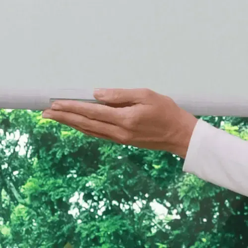 Hand touches a white roller shade in front of a green, leafy background.