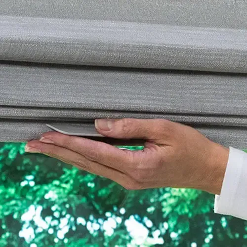 Hand adjusting a gray Roman shade in front of a window with green foliage.