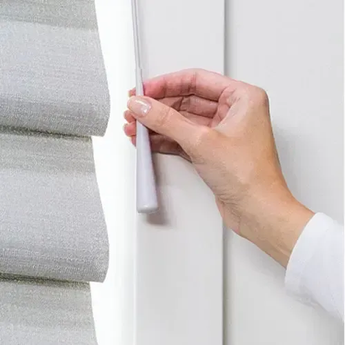 Hand pulling cord to adjust a light gray, pleated Roman shade. White wall.