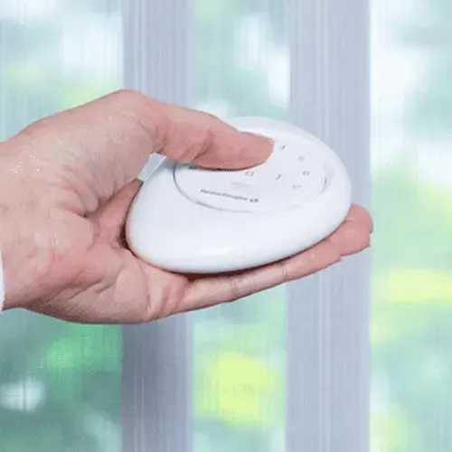 Hand holding a white, oval-shaped remote with numbered buttons, in front of a window.
