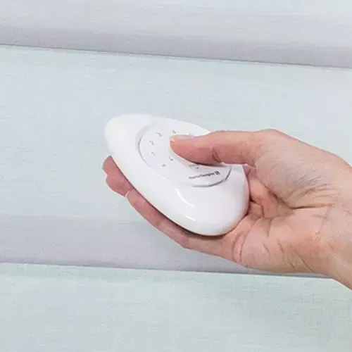 Hand holding white remote control, likely for blinds, against a light background.