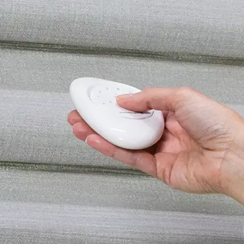 Hand holding a white remote, controlling light-colored window blinds.