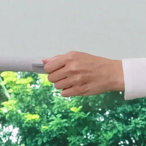 Hand pulls down a window shade. Green foliage visible outside the window.