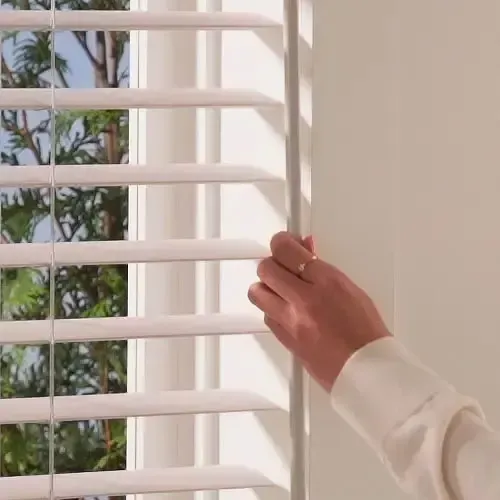 Hand adjusting white window blinds, natural light visible.