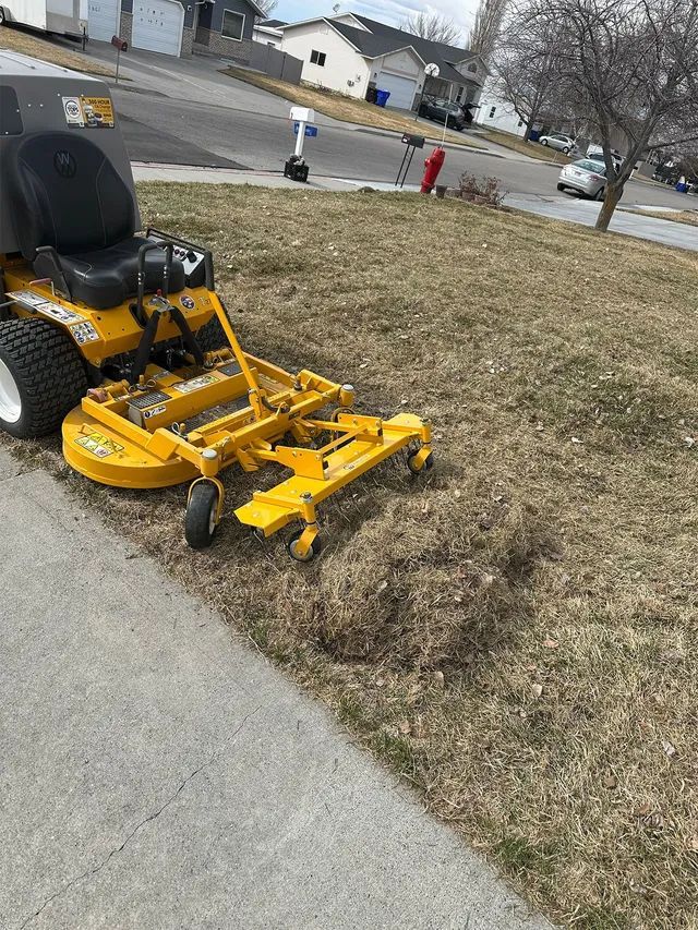 Yellow riding lawn mower on a grassy roadside lawn near a sidewalk and suburban street.