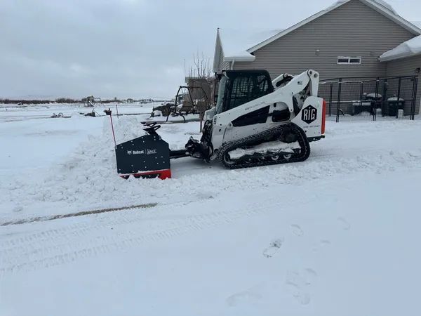 White skid-steer loader with snowplow in snowy driveway beside a house and garage