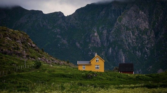 Yellow house and small brown house sit on green hillside, mountains in the background.
