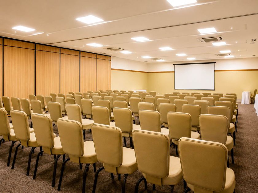Sala de conferências vazia com fileiras de cadeiras bege e uma tela de projeção na frente.