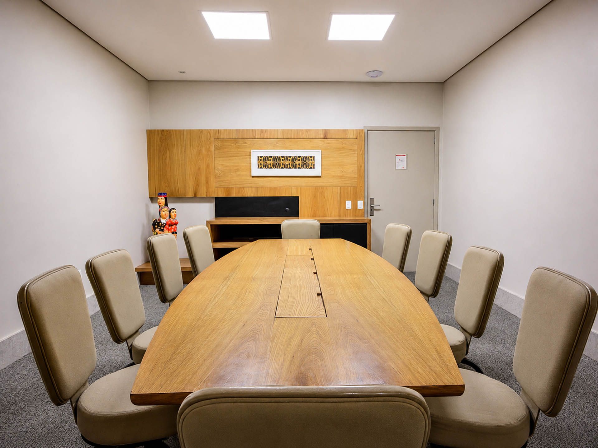 Sala de conferências vazia com uma longa mesa de madeira, cadeiras bege e um quadro na parede do fundo.