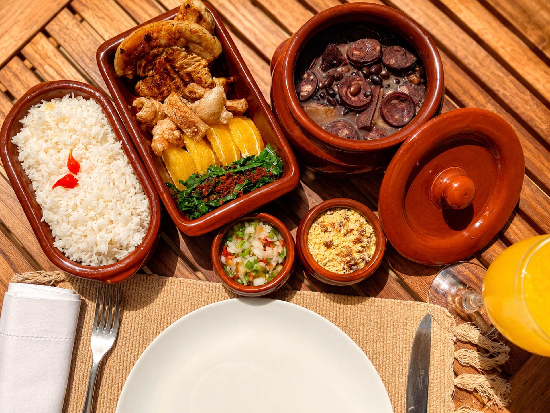 Pratos variados do Sudeste Asiático em panelas de barro, acompanhados de arroz e bebidas, sobre uma esteira de palha.