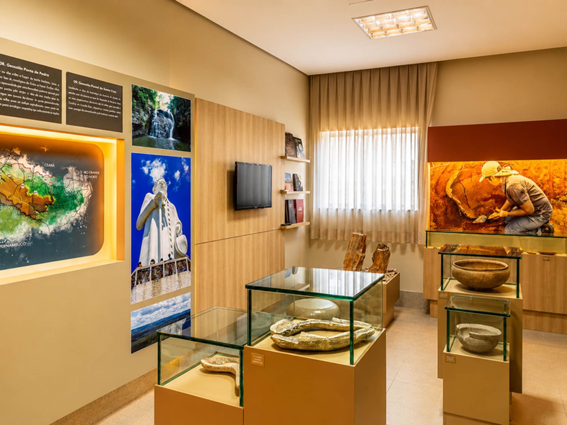 Sala de exposições do museu com vitrines de vidro, painéis de parede e obras de arte coloridas ao redor de uma janela.
