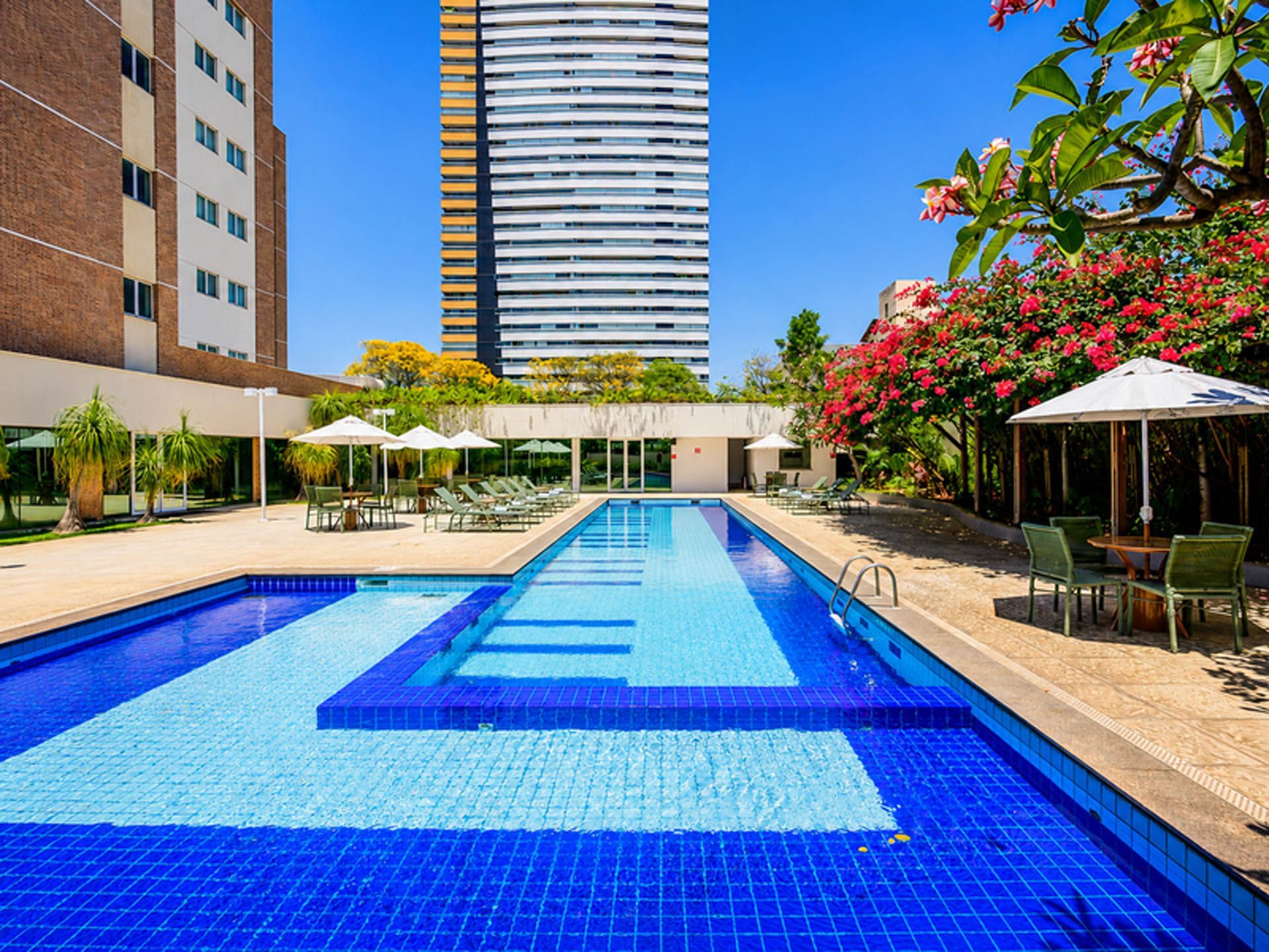 Piscina externa de hotel com água azul, espreguiçadeiras, guarda-sóis e prédios altos ao fundo.