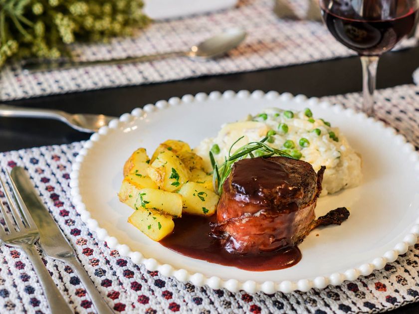 Bife servido com batatas e purê de batatas ao lado de uma taça de vinho tinto em uma mesa de jantar.