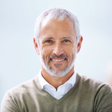 Smiling man with gray hair and beard in a white shirt and olive sweater against a light background