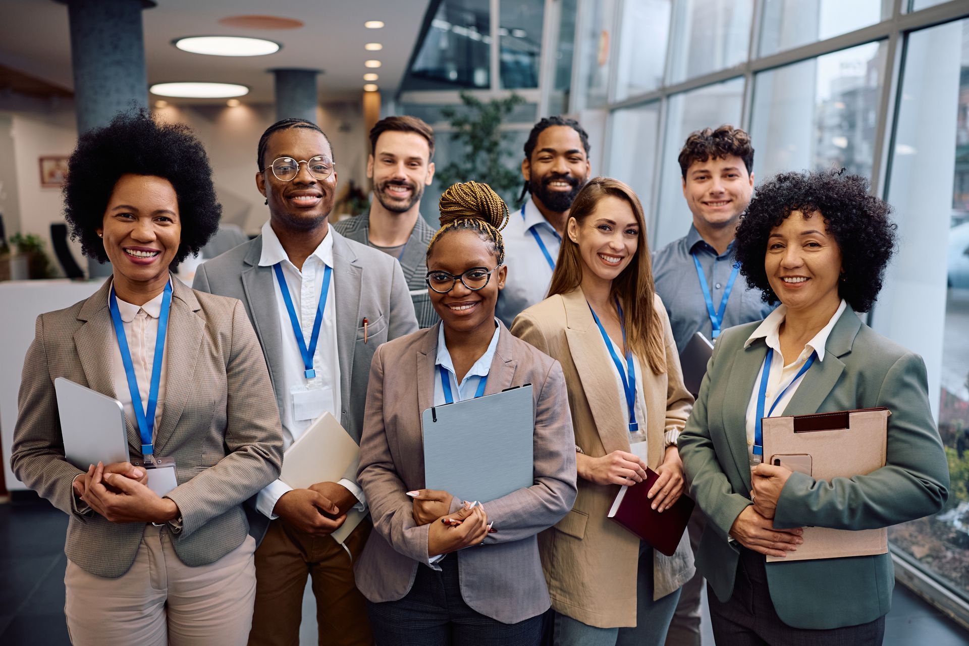Group of smiling coworkers wearing lanyards, standing in an office lobby and holding notebooks and folders.