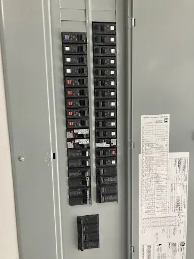 Electrical panel with rows of black circuit breakers in a gray metal enclosure. A paper label is visible.