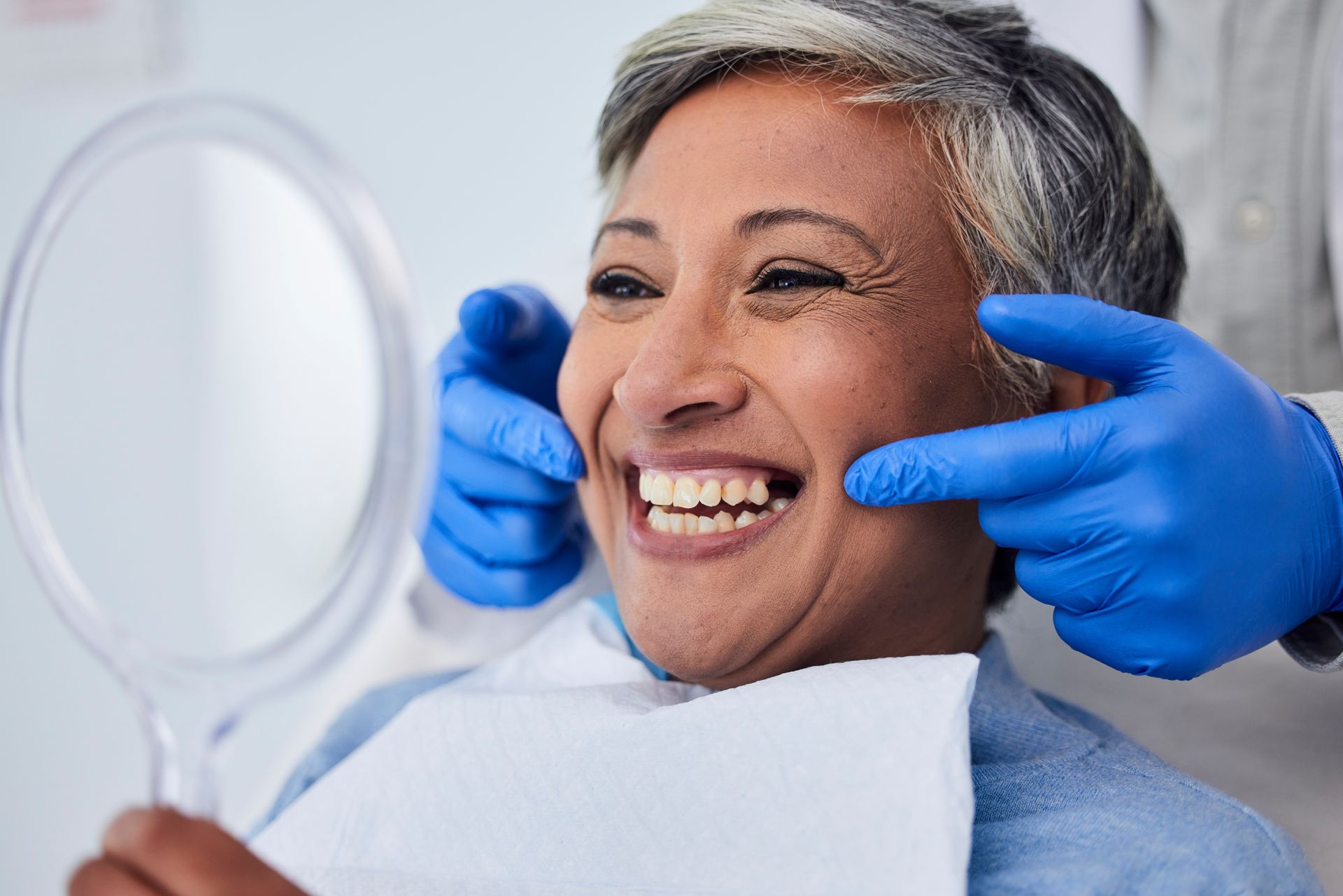 A female patient smiling while holding a hand mirror. A dentist is behind her, pointing at her mouth