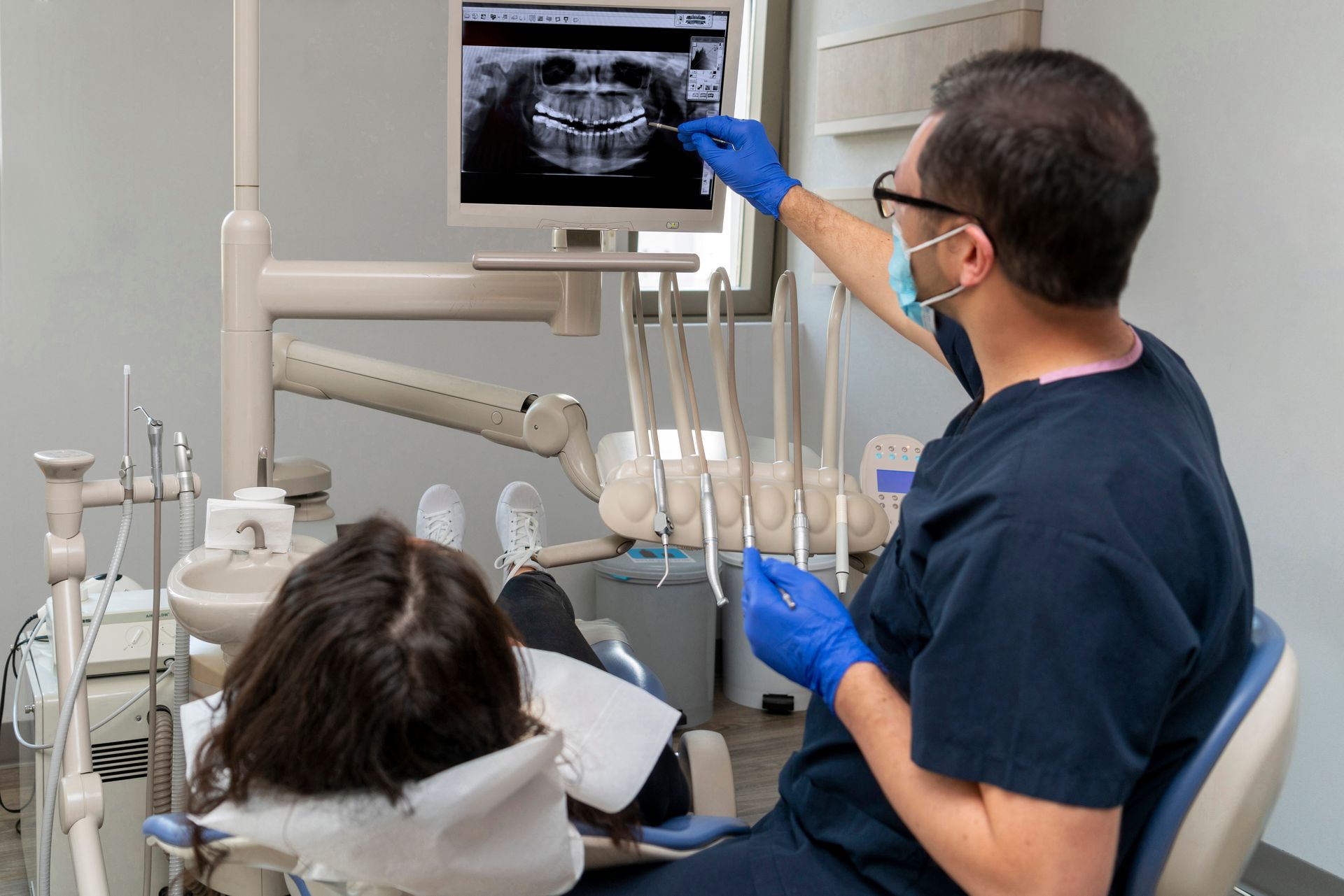 Dentist pointing at x-ray on screen, explaining procedure to patient in dental chair.