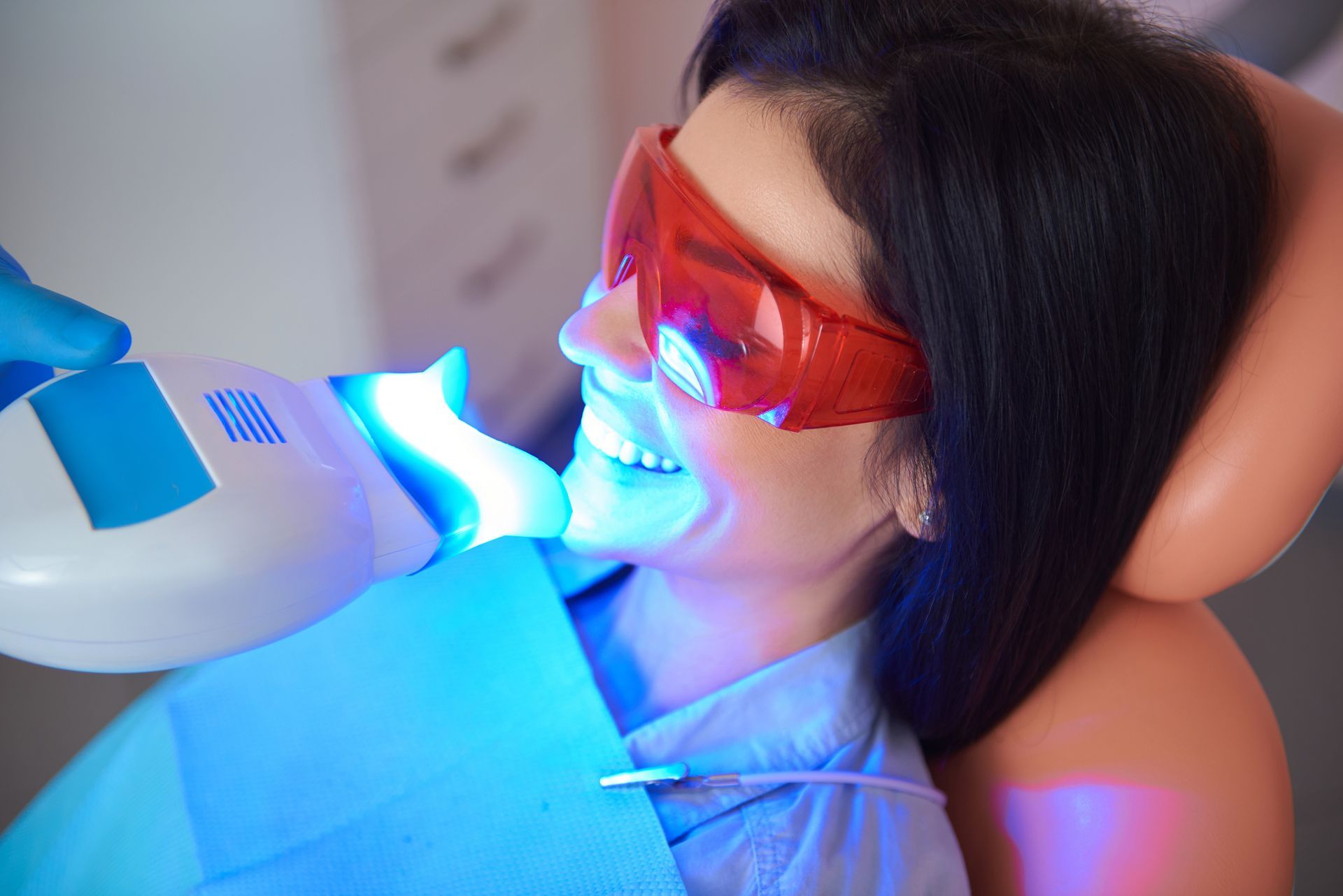 Woman in chair receiving teeth whitening treatment with blue light, wearing protective glasses.