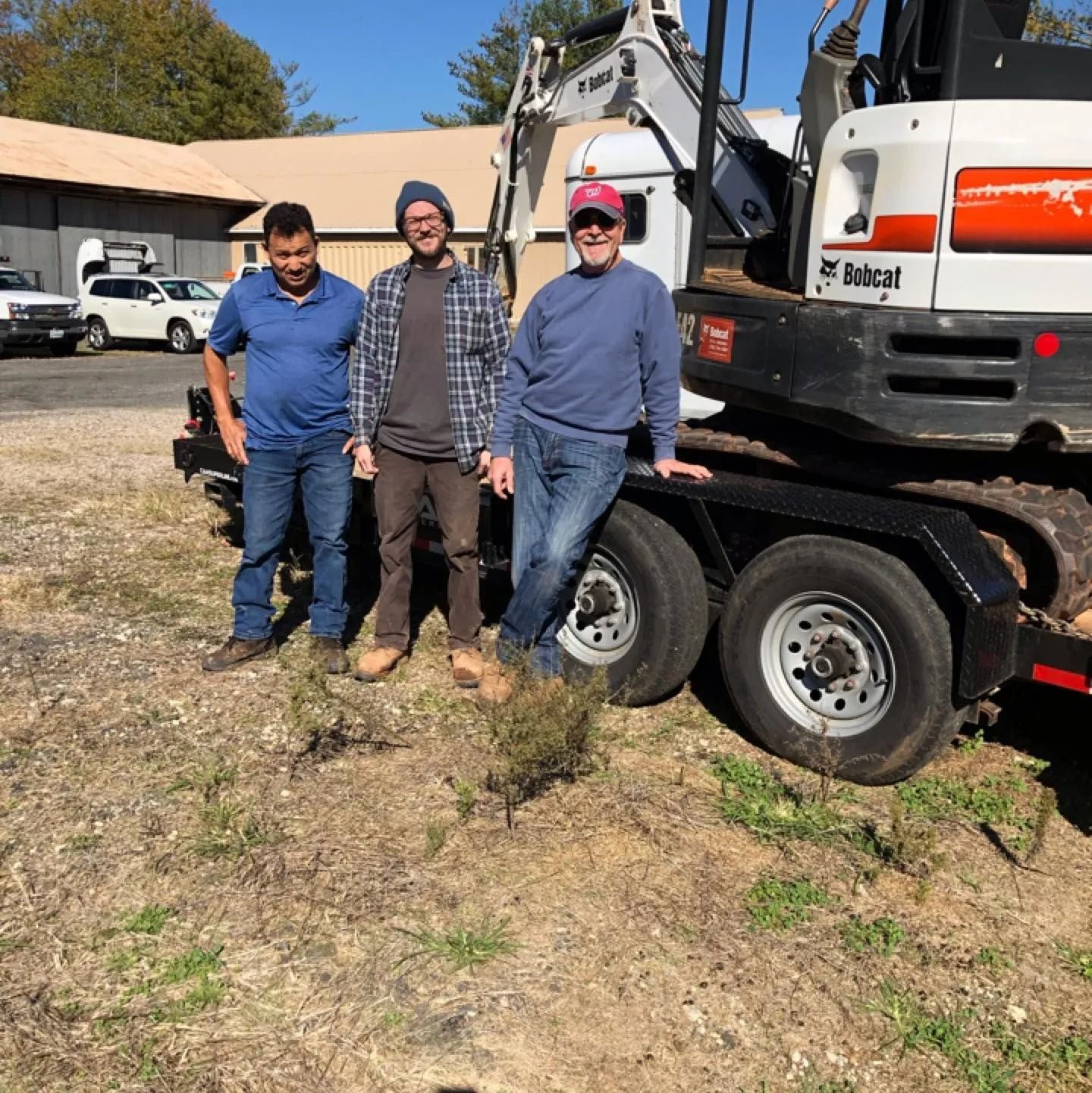 Three men stand beside a Bobcat excavator on a trailer outdoors on a sunny day.