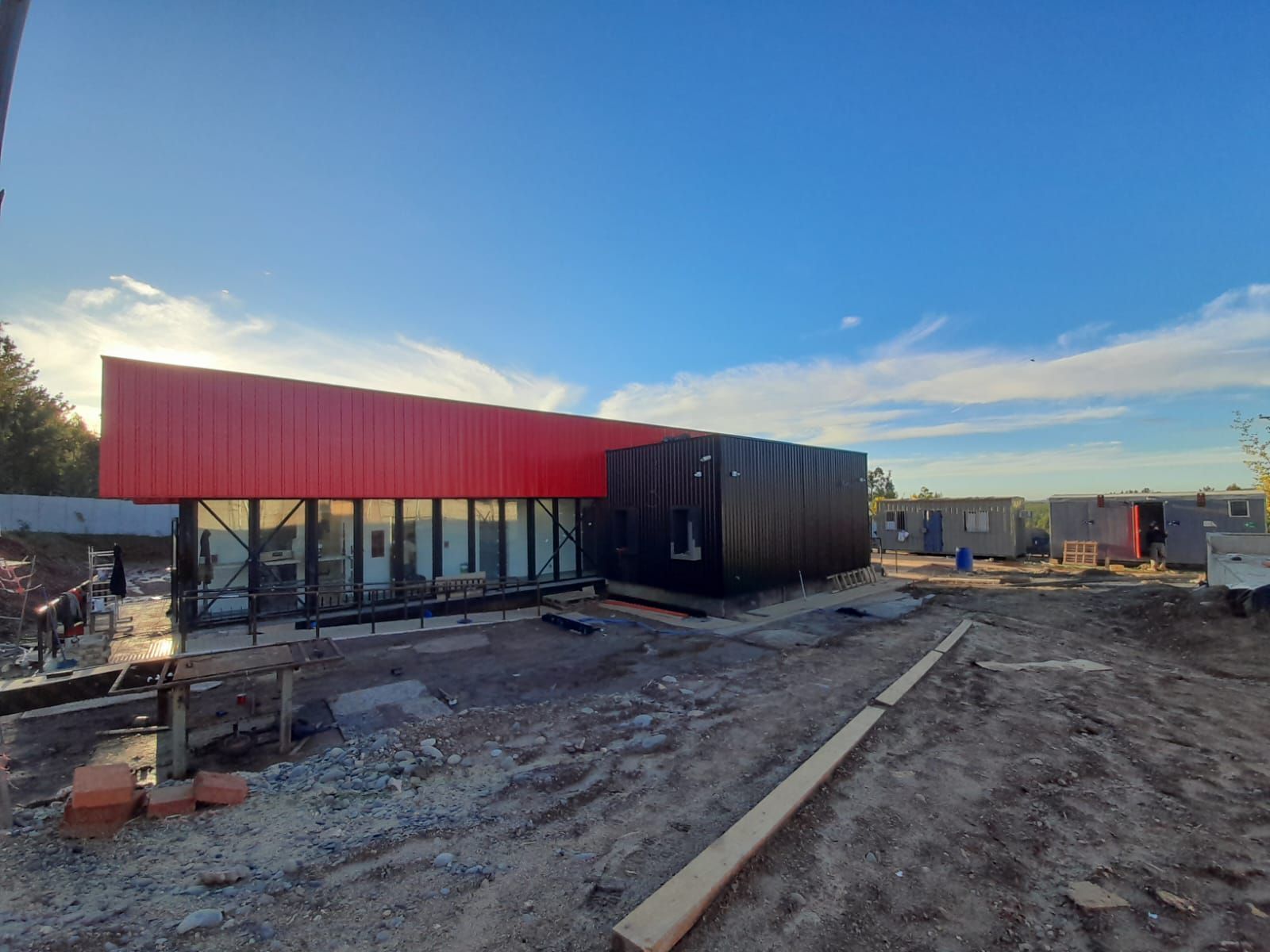 Edificio en construcción con techo rojo, sección negra y grandes ventanales, contra un cielo azul.