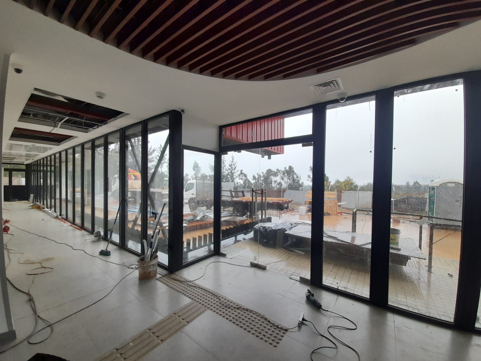 Interior de un edificio en construcción, largo pasillo con grandes ventanales que dan a una escena exterior húmeda.