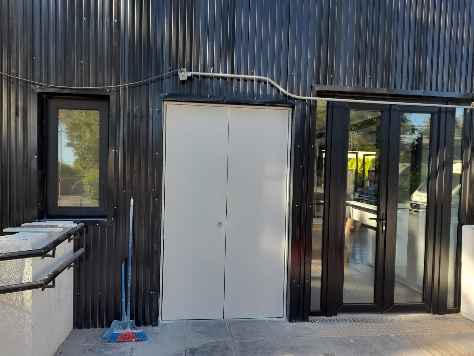 Exterior de edificio negro con una puerta doble blanca, una ventana y una puerta de vidrio; una escoba apoyada en la pared.
