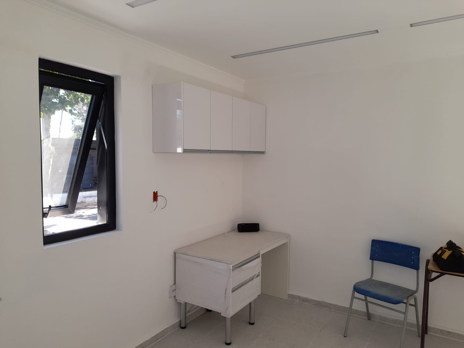 Una oficina pequeña y vacía con un escritorio, armarios, una silla y una ventana. Las paredes y los armarios son blancos.